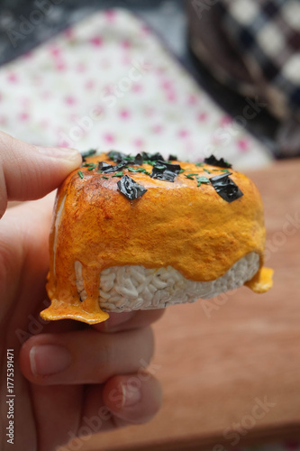 a hand holding 1 on 1 replica of japanese onigiri topped with mentai sauce and seaweed on wooden background