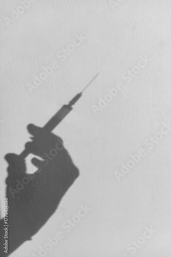 Shadow of hand holding syringe against light wall, thin needle visible, minimal monochrome medical concept