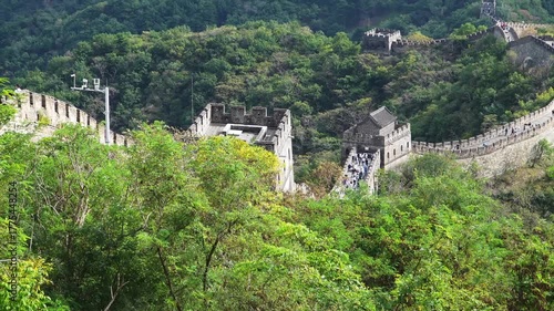 Great Wall of China winding through lush green mountains with tourists. Ancient stone fortification with watchtowers amid dense forest. Historic landmark and World Heritage Site summer view.
