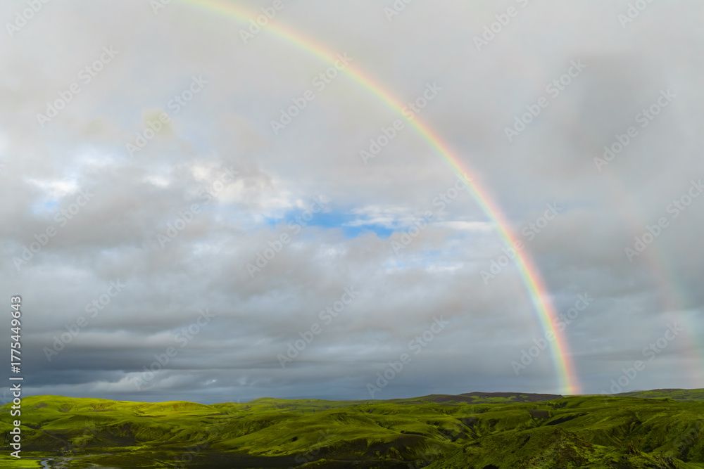 Naklejka premium A bright rainbow arcs above moss green lava fields near Blue Lagoon in southwest Iceland. Dark basalt patches and a winding stream sit under clouds breaking to blue.
