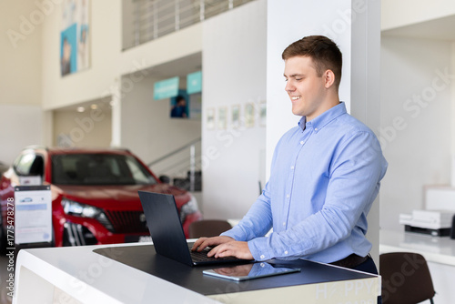Happy car salesman working on laptop in dealership office. Young professional checking customer data and vehicle information inside modern automotive showroom