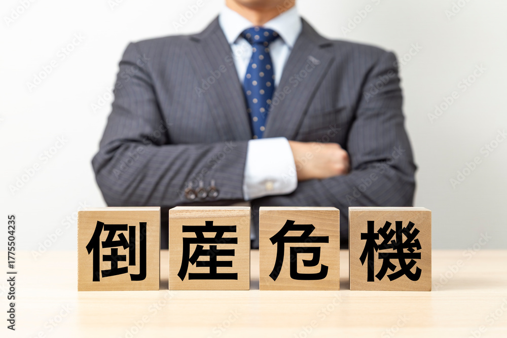 Obraz premium 「倒産危機」と書かれたブロックと腕組みをするビジネスマン