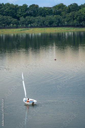 A man in an orange life jacket sails a small boat in a pond in a park.