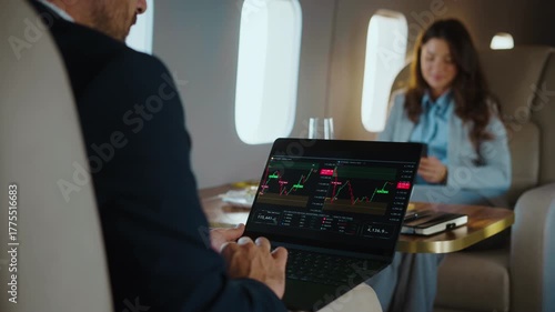 Focused businessman working on a laptop, checking stock market charts and financial data while traveling on a luxurious private jet with a female colleague sitting in front of him
