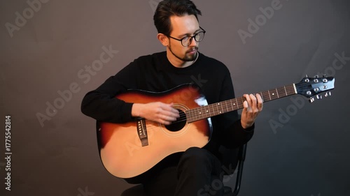 A handsome young brunet man playing guitar while sitting on a chair in a dark studio

