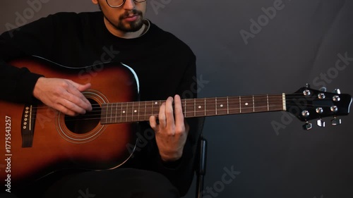 A handsome young brunet man playing guitar while sitting on a chair in a dark studio

