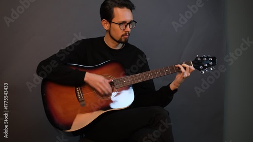 A handsome young brunet man playing guitar while sitting on a chair in a dark studio

