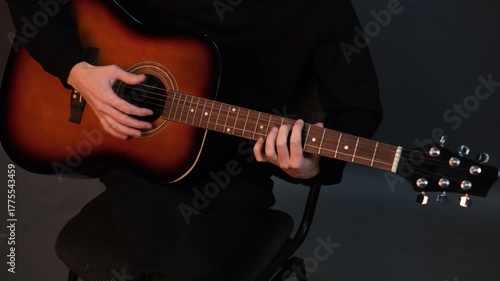 A handsome young brunet man playing guitar while sitting on a chair in a dark studio

