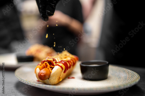 A gourmet hot dog is prepared by a chef in black gloves, with ketchup and mustard drizzled on a sausage in a bun, and a topping sprinkled over it.