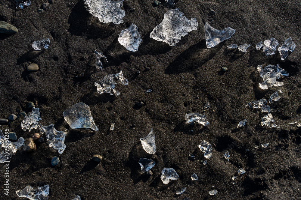 Fototapeta premium Glistening shards of ice rest on the dark, volcanic sand, a stark contrast.