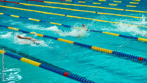 Swimmers competing in a race at an indoor pool with blue and yellow lane dividers. Action shot capturing splashes and movement in clear turquoise water