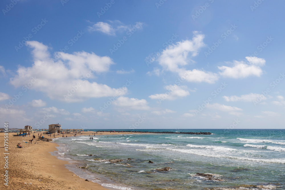 Fototapeta premium Haifa, Israel, A beach on a clear day, with moderate waves breaking on the shore near a small rock formation, people enjoying the sand, and a small complex of buildings with thatched umbrellas