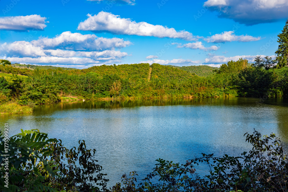 Fototapeta premium 2025-10-02 CALM LAKE FRAMED AND SURROUNDED BY GREEN FOLIAGE AND A ROLLING HILL IN THE BACKGROUND AND A NICE CLOUDY SKY NEAR SIENA IN THE TUSCUNY REGION OF ITALY