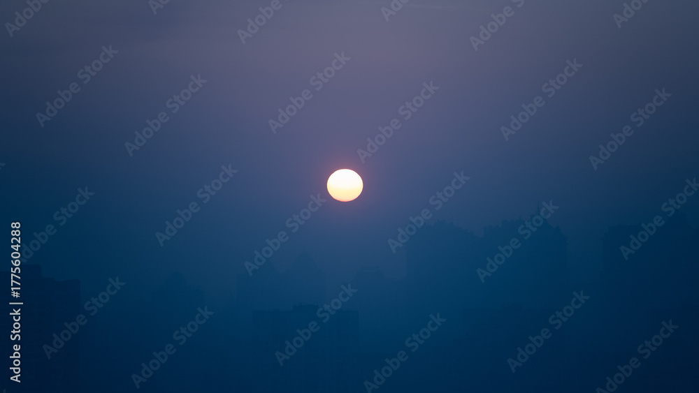 Obraz premium A warm sun sets in the evening, surrounded by a hazy sky that provides a tranquil backdrop. The city skyline is barely visible, enhancing the peaceful scene