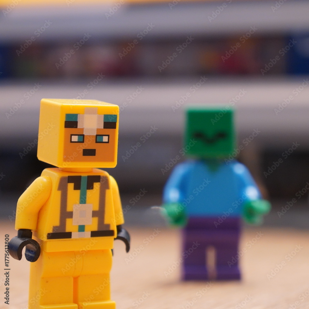 Naklejka premium A LEGO minifigure in a protective yellow suit stands beside a green-skinned LEGO zombie from the Minecraft series, showcasing popular toy figures and imaginative play