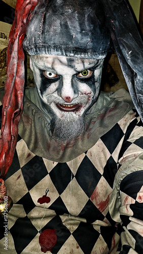A man dressed as an evil jester or clown, with different colored contact lenses, covered in blood stains.