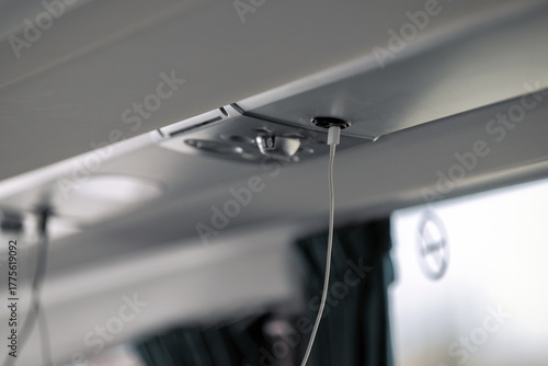 A close-up shows a white charging cable plugged into a black USB port located in the overhead panel of a modern coach bus, highlighting passenger technology.