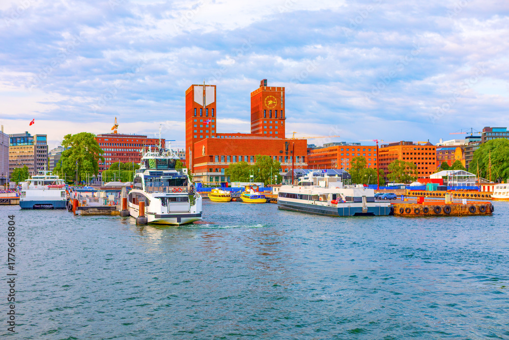 Fototapeta premium Oslo City Hall prominent brick building with two towers, overlooking the harbor in Oslo, Norway, boats docked in foreground. Cloudy sky casting soft light on city waterfront and surrounding buildings