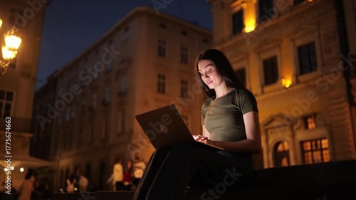 Young woman working on a laptop outdoors at night, illuminated by screen light, sitting on city steps surrounded by warm street lamps and evening crowds