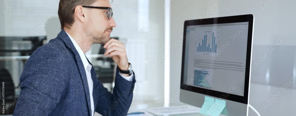 Fototapeta premium Businessman in a blue jacket analyzing financial data charts on computer, making important business decisions in modern office. Business people concept