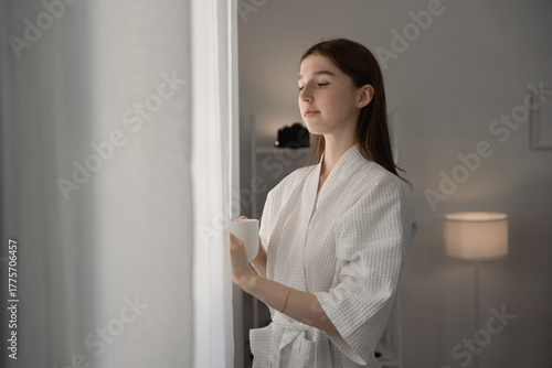 Free Download Young woman in white bathrobe standing by window with cup of coffee, enjoying peaceful morning light at home. Perfect concept of relaxation, slow living. woman, bathrobe, coffee, morning. cozy minimal - GFXLoad