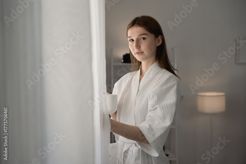 Free Download Young woman in white bathrobe standing by window with cup of coffee, enjoying peaceful morning light at home. Perfect concept of relaxation, slow living. woman, bathrobe, coffee, morning. cozy minimal - GFXLoad