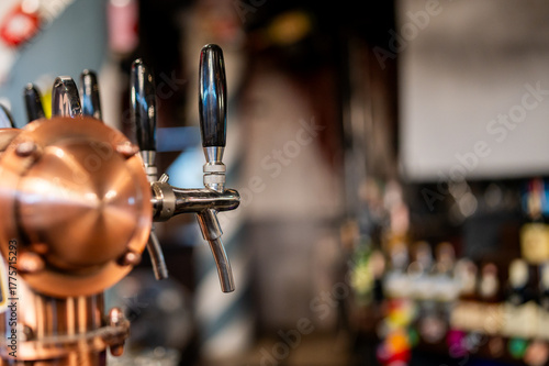 A close-up, selective focus shot of a row of shiny chrome and copper beer taps on a bar counter with a blurred background of a pub.