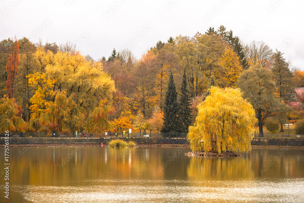 Naklejka premium This tranquil pond in Kudowa Zdroj features vibrant autumn colors with shades of orange and yellow. Visitors stroll along the water's edge, enjoying the peaceful fall atmosphere.