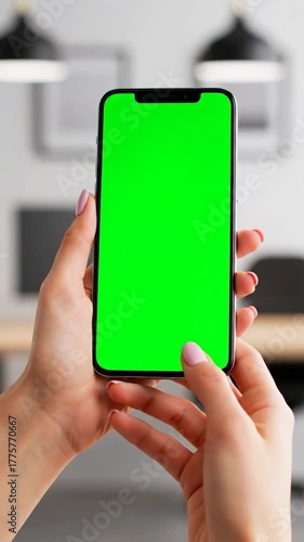 Close-up of hands holding a smartphone with a green screen in a blurred interior
