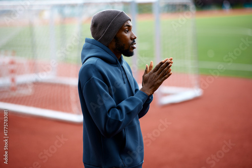 Focused black male athlete wearing a hoodie and a beanie rubs his hands together to warm up or concentrate before starting a workout on an outdoor sports field