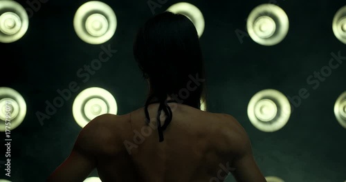 bikini fitness competitor stands with their back to the camera showcasing their physique under vibrant stage lights. The atmosphere reflects excitement in a fitness competition.