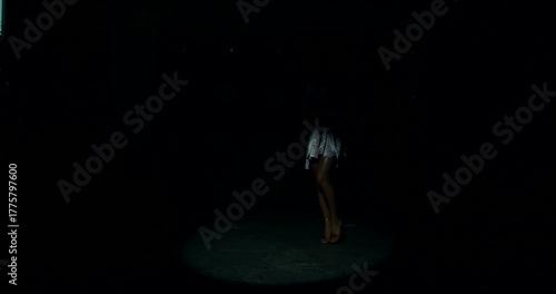 Under a beam of light a fitness bikini model steps forward onstage revealing a stunning swimsuit. The backlight highlights her form as she prepares for the event.