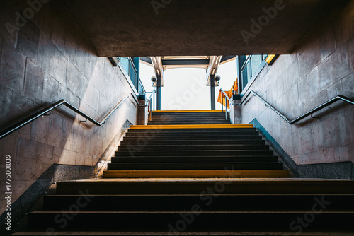 View from the bottom of a stairway leading up to street level in an urban subway or underpass. Sunlight brightens the exit framed by concrete walls, metal railings, and a hint of sky above