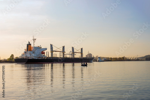 A large cargo ship is pictured on calm water. In the foreground, a small fishing boat with a few people on board floats on the water.