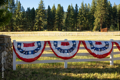 Red, white, and blue patriotic bunting with white stars on the blue section, shaped in semicircles and draped along a fence for Fourth of July decoration. A slice of Americana in a rural area.