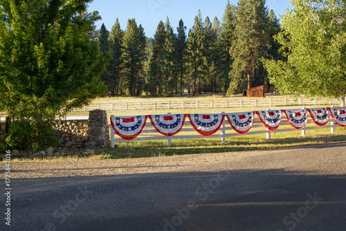 Red, white, and blue patriotic bunting with white stars on the blue section, shaped in semicircles and draped along a fence for Fourth of July decoration. A slice of Americana in a rural area.