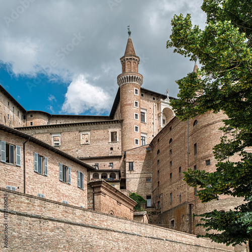 The majestic Palazzo Ducale in Urbino, Marche, Italy, seen from below, is rising from the other buildings. Renaissance brick architecture with a tall tower against a cloudy sky.