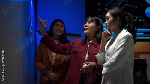 Three women are standing in front of a whiteboard, writing on it with markers. They seem to be working together on a project or discussing ideas. Scene is collaborative and focused