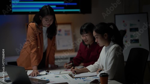 Three women are sitting at a table with papers and a laptop. They are discussing something important. Scene is serious and focused