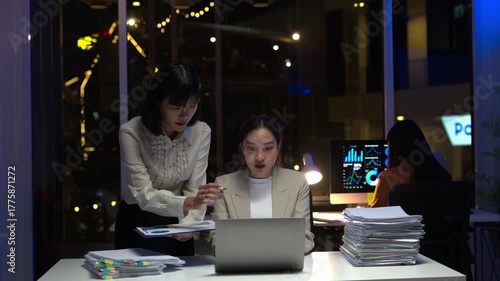 Two women are sitting at a desk with a laptop and a clipboard. One of the women is writing on the clipboard