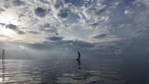 A sailing boat in the calm and reflecting sea with the sun breaking through the clouds after a storm in the Aegean Sea.