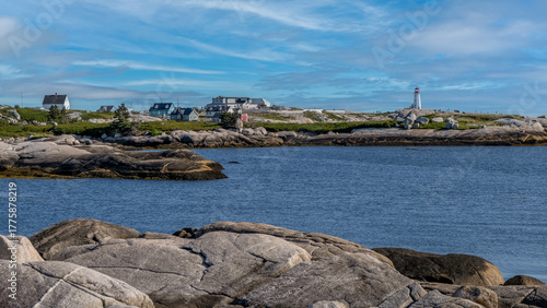 Slika na platnu Rocky Atlantic coastline at Cape Breton, Nova Scotia, with blue ocean, seaside houses, and a distant white lighthouse under a bright sky