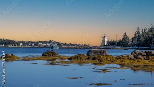 Photos Calm coastal inlet at Cape Breton, Nova Scotia, with low tide rocks, a moored fishing boat, and a white lighthouse at sunset under a clear sky