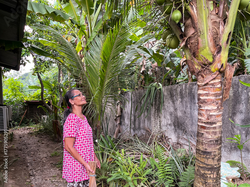 An elderly Asian woman stands and looks at coconuts on palm trees