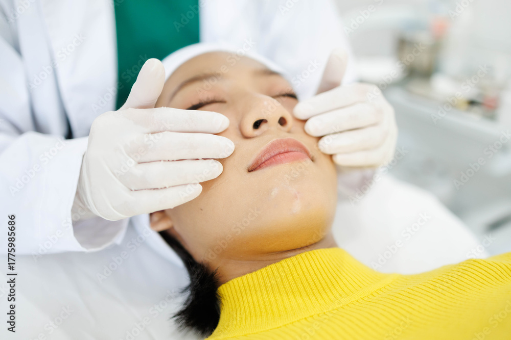 Obraz premium A woman is getting her face massaged by a man in a white lab coat. The woman is wearing a yellow sweater and the man is wearing gloves. The scene is set in a medical setting