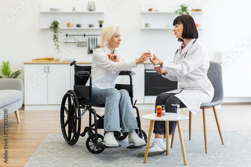 A female doctor is giving medication to an elderly woman in a wheelchair, discussing treatment in a home setting.