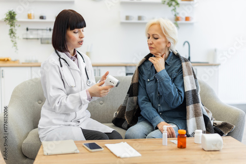 A female doctor is checking an elderly patient's temperature using a digital thermometer while the patient sits on a couch.