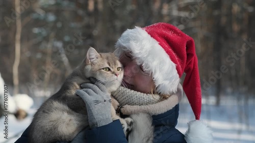 A joyful woman in a Santa's hat plays with a furry cat in the woods with a snowy trees on the background. Happy young girl walking with her kitten in the winter park enjoying christmas vacations.
