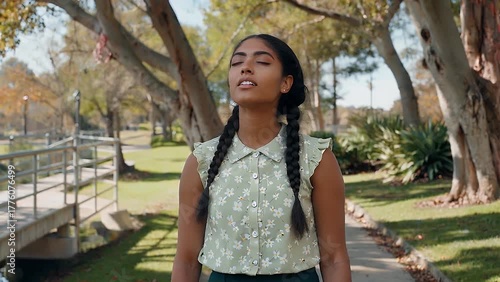 A youthful woman of a specific nationality enjoying a moment of tranquility with closed eyes while inhaling fresh outdoor air in a green park setting