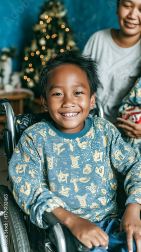 A young boy in a wheelchair smiles brightly while sitting indoors. A caregiver and a decorated Christmas tree are visible in the warmly lit background.
